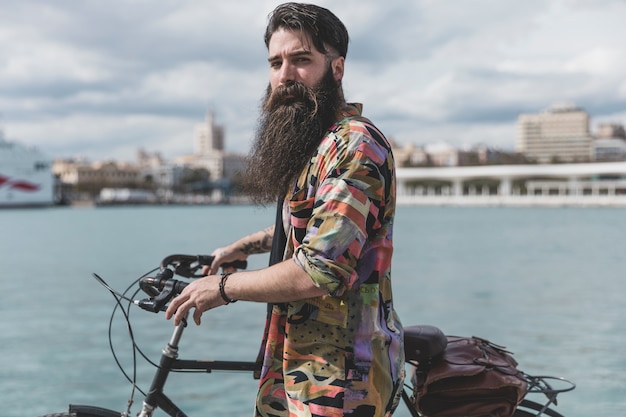 Langer bärtiger junger Mann, der mit Fahrrad nahe der Küste steht