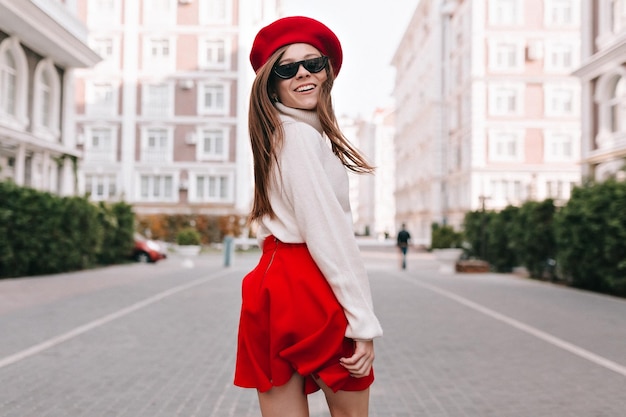 Langhaarige attraktive Frau in stylischem rotem Baskenrock und Brille lächelt breit und posiert auf Stadthintergrund