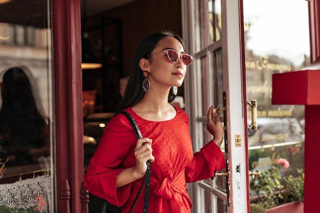 Langhaarige brünette Frau in roter Sonnenbrille und trendigem hellem Kleid schaut weg und öffnet Cafétür