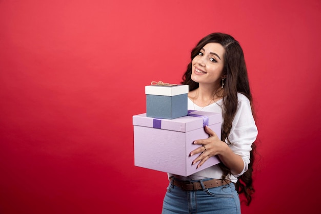 Langhaarige Frau, die mit Geschenkboxen auf rotem Hintergrund steht. Foto in hoher Qualität