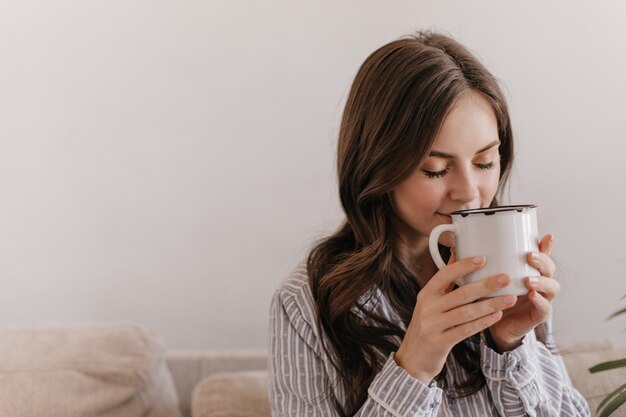 Langhaarige Frau im blauen Pyjama, die das Aroma des Tees genießt