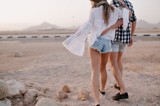 Langhaarige frau mit mann in trendigen jeansshorts geht am frühen morgen in einer umarmung in der nähe der autobahn spazieren. stilvolles paar umarmt sich und genießt einen schönen blick auf die wüste an einem date am sommerabend.