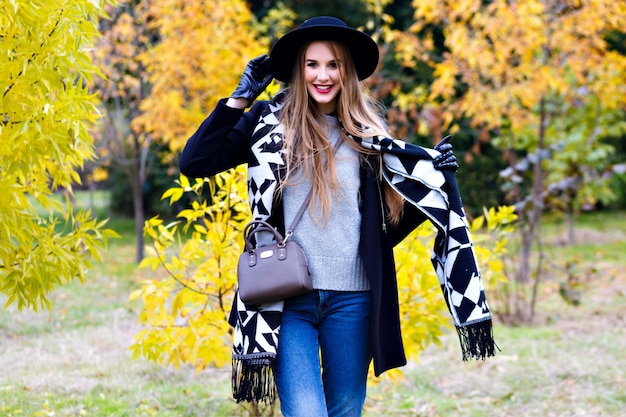 Langhaariges Mädchen trägt Jeans, die mit ihrem Schal im Herbstpark spielen. Attraktive junge Frau im stilvollen schwarzen Hut, der Zeit im Wald verbringt und lacht.
