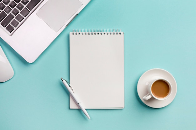 Laptop mit gewundenem Notizblock, Maus, Kaffeetasse und Stift auf blauem Schreibtisch