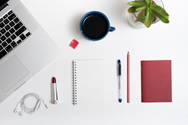 Laptop; Teetasse; Kopfhörer; Lippenstift und Büromaterial auf weißem Schreibtisch