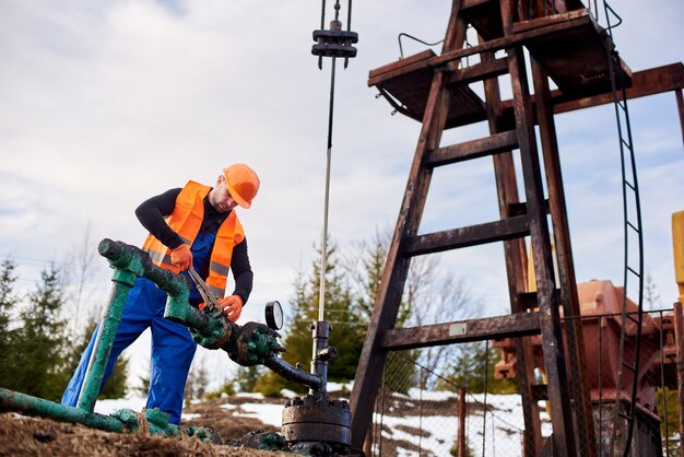 Ölarbeiter in orangefarbener Uniform und Helm, der mit einer Rohrzange in der Nähe eines Ölpumpenhebers arbeitet