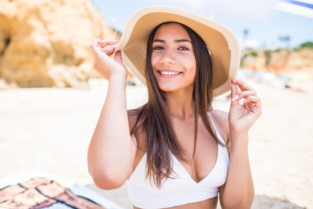 Lateinische Frau der jungen Schönheit im Bikini und im Strohhut, die unter Sonnenschirm am Strand nahe der Seeküste sitzen.