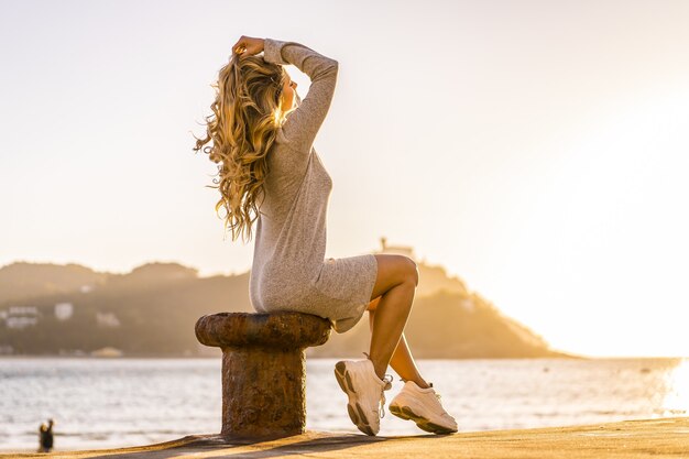 Lateinische Frau mit blonden Haaren, die bei Sonnenuntergang am Meer an der Küste sitzt