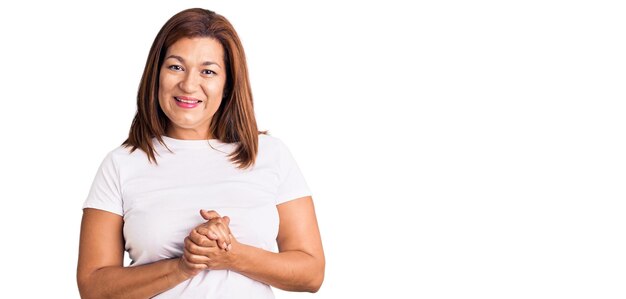 Lateinische Frau mittleren Alters, die ein lässiges weißes T-Shirt mit zusammengelegten Händen und gekreuzten Fingern trägt und entspannt und fröhlich lächelt. Erfolg und Optimismus