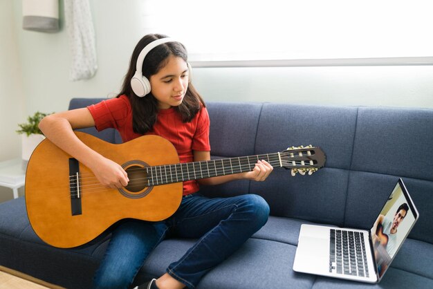 Lateinisches Mädchen mit Kopfhörern, das ihren Online-Musikunterricht hört. Künstlerisches Kind, das Akustikgitarre spielt und die Akkorde lernt