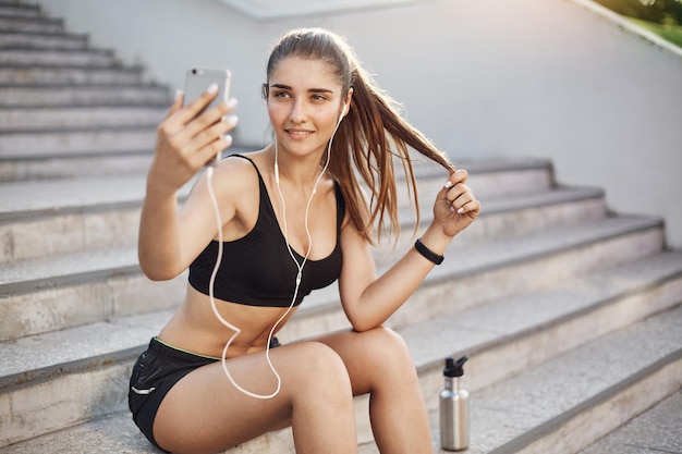 Laufende Tranerin, die ein Selfie mit einem Smartphone macht, das Rockmusik in ihren weißen kabelgebundenen Kopfhörern hört und alle ihre Freundinnen vor Neid auf ihren perfekten Körper rosa macht