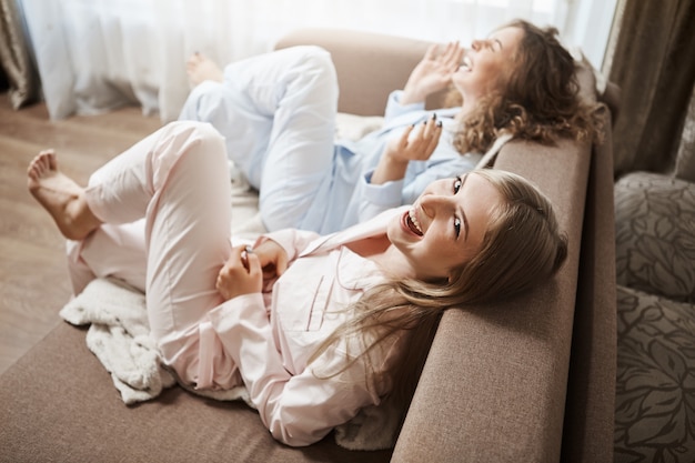 Lazy-Bones tun nichts und genießen gemeinsam die Freizeit. Oberwinkelaufnahme von niedlichen Frauen, die in Nachtwäsche auf dem Sofa sitzen und laut lachen, über unangenehme Momente im Büro sprechen und Wochenenden verbringen