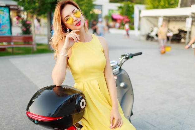 Lebensstil-Außenporträt der stilvollen Frau im gelben Weinlesekleid sitzen auf schwarzem Elektromotorrad.