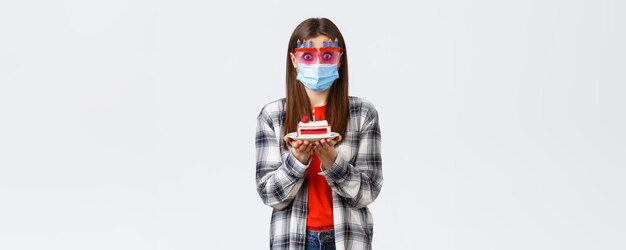 Lebensstil des Coronavirus-Ausbruchs während der sozialen Distanzierung und des Feiertagsfeierkonzepts Überrascht oder überfallenes süßes Mädchen in Brille und medizinischer Maske mit Geburtstagstorte verwirrt, wie Kerze bläst