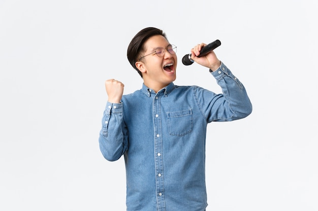 Lebensstil, Freizeit und Menschenkonzept. Sorgloser glücklicher asiatischer Mann, der genießt, beim Karaoke zu singen, Mikrofon und Faustpumpe im Entzücken hält und über weißer Wand auftritt