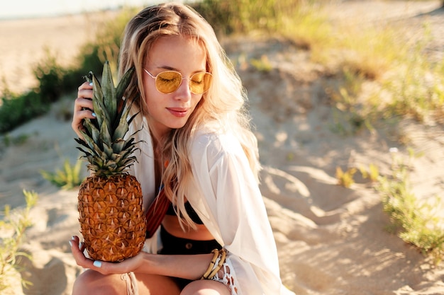 Lebensstil im Freienbild der lachenden hübschen Frau mit saftiger Ananas, die am sonnigen Strand entspannt. Trendiges Sommeroutfit