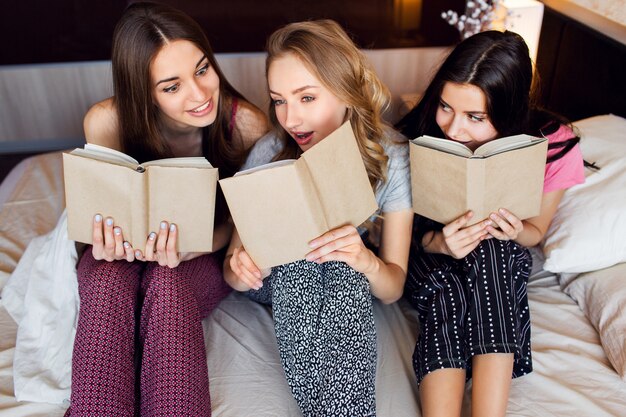 Lebensstilbild von drei besten Freunden im Pyjama, die Bücher lesen, studieren und Gespräche im Schlafzimmer führen. Gruppe von Studenten, die zusammen zu Hause Hausaufgaben machen. Gemütliche warme Farben. Emotionale Gesichter.