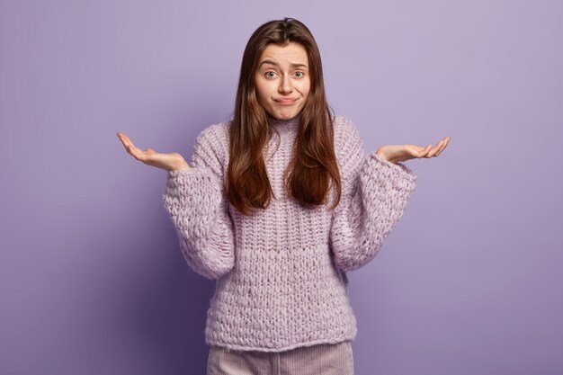 Lebenswahrnehmungskonzept. Die ahnungslose europäische Frau zuckt mit den Schultern, trifft eine Entscheidung, trägt einen violetten Pullover, hat einen zögernden Gesichtsausdruck, steht und gestikuliert isoliert über der lila Wand