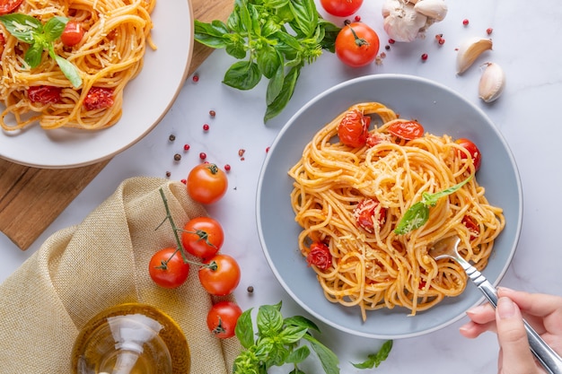 Leckere appetitliche klassische italienische Spaghetti-Nudeln mit Tomatensauce, Käseparmesan und Basilikum auf dem Teller und Zutaten zum Kochen von Nudeln auf weißem Marmortisch. Flache Draufsicht Kopienraum.
