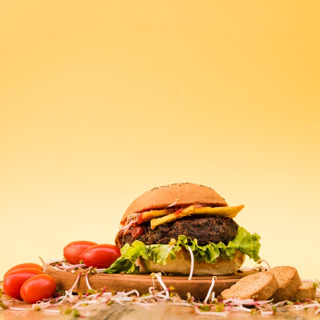 Leckere Hamburger mit Kirschtomaten; Sprossen und Brotscheiben auf Schneidebrett vor gelbem Hintergrund