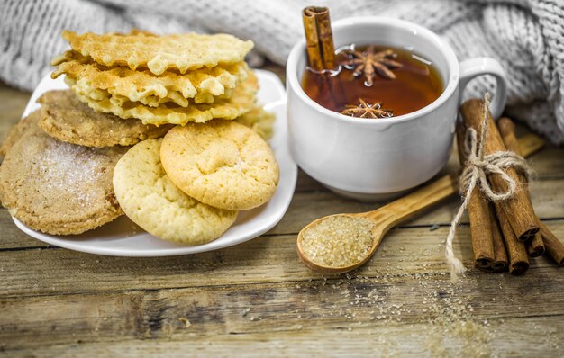 leckere Kekse und eine Tasse heißen Tee mit einer Zimtstange und einem Löffel braunem Zucker auf Holz