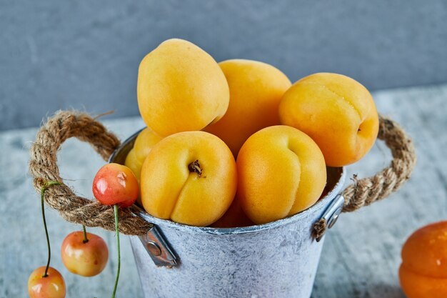 Leckere reife Aprikosen im Eiseneimer mit Kirschen auf Marmoroberfläche