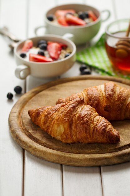 Leckere schöne Croissants auf Holzbrett. Traditionelles kontinentales Frühstück. Granola mit Früchten und Honig auf Hintergrund.