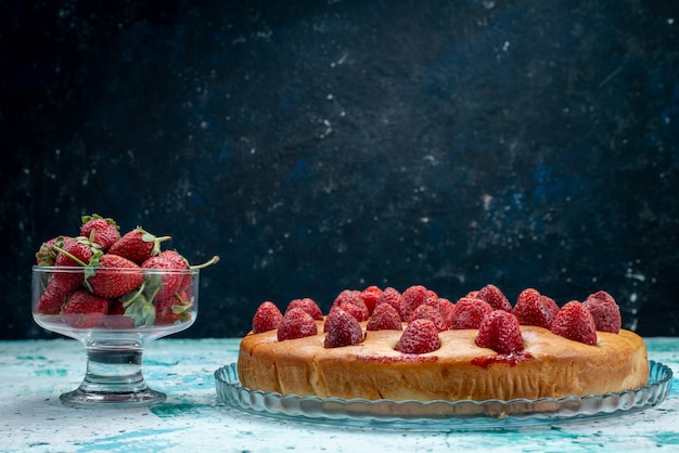 leckerer Erdbeerkuchen rund geformt mit Früchten und zusammen mit frischen roten Erdbeeren auf hellblauem Schreibtisch, Kuchenteig Keksfruchtbeere