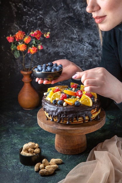 Leckerer Schokoladenkuchen der Vorderansicht, der mit frischem Obst von Frau auf dunkler Wand verziert wird