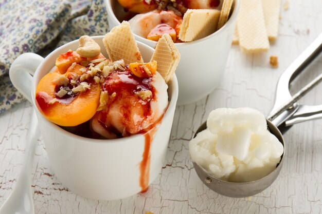 Leckeres frisches kaltes appetitliches Vanilleeis mit Nüssen, Aprikosen, Waffeln und Sirup auf weißem Tisch mit Zutaten zum Dessert. Nahansicht.