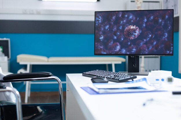 Leere helle Arztpraxis mit Computer auf Schreibtischtisch mit Cornavirus-Illustration auf dem Bildschirm während der globalen Pandemie covid19. Krankenzimmer mit professionellen Werkzeugen ausgestattet. Virenbild