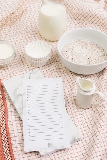 Leere Liste im Tagebuch mit Mehl; Milchglas und Formen über Tuchhintergrund