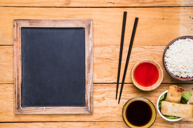 Leere Tafel neben den Essstäbchen; Frühlingsrollen; Reis und Saucen auf Schreibtisch aus Holz