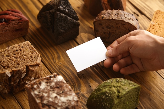 Leere Visitenkarte des professionellen handwerklichen Bäckers präsentiert in der Hand in der Mitte vieler gemischter alternativer gebackener exotischer Brotproben über hölzernem rustikalem Tisch