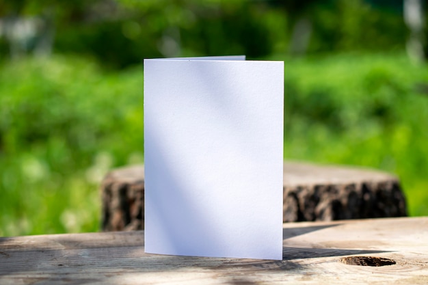 Leere zweifache weiße Karte, die auf Holzschreibtisch im Freien mit Blumenschatten und unscharfem Naturhintergrund steht