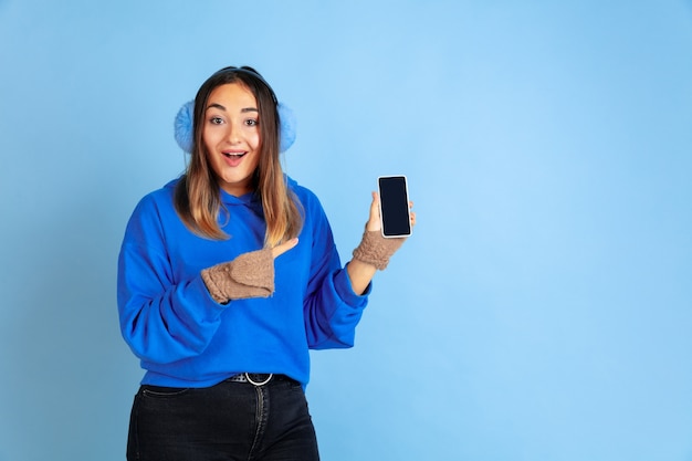 Leeren Bildschirm anzeigen. Porträt der kaukasischen Frau auf blauem Studiohintergrund. Schönes weibliches Modell in warmer Kleidung. Konzept der menschlichen Emotionen, Gesichtsausdruck, Verkauf, Anzeige. Winterstimmung.