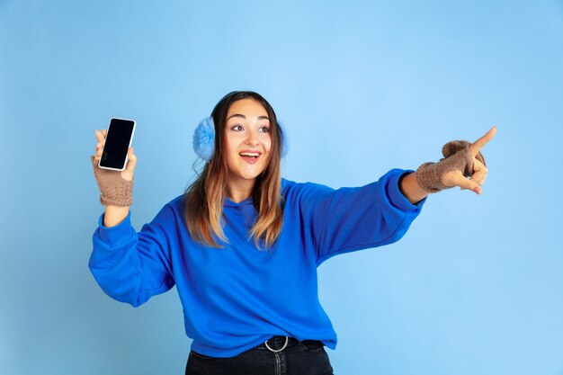 Leeren Bildschirm anzeigen. Porträt der kaukasischen Frau auf blauem Studiohintergrund. Schönes weibliches Modell in warmer Kleidung. Konzept der menschlichen Emotionen, Gesichtsausdruck, Verkauf, Anzeige. Winterstimmung.