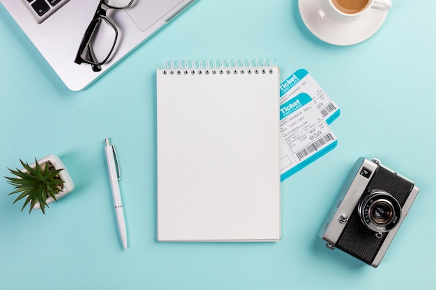 Leerer gewundener Notizblock mit den Flugtickets umgeben mit Laptop, Brillen, Stift, Kamera, Kaffeetasse auf blauem Schreibtisch
