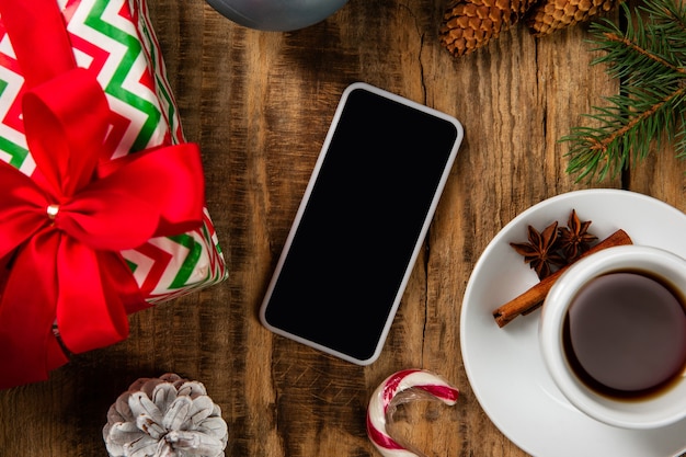 Leerer leerer Bildschirm des Smartphones an der Holzwand mit bunter Feiertagsdekoration, Tee und Geschenken.
