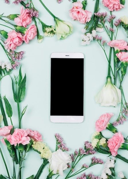Leeres Smartphone mit frischem Limonium umgeben; Nelken und Eustoma Blumen vor blauem Hintergrund