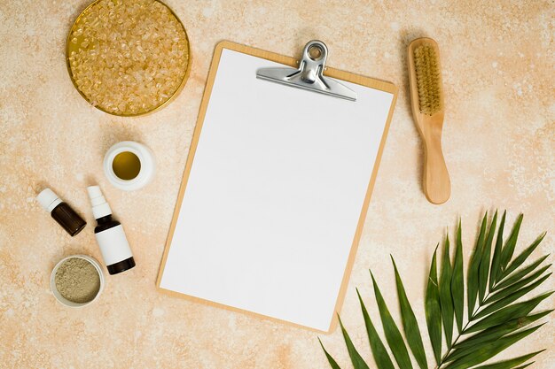 Leeres weißes Papier in der Zwischenablage, umgeben von Rhassoul-Ton; Salz; Honig; essentielle Öle; Pinsel und Palmblätter