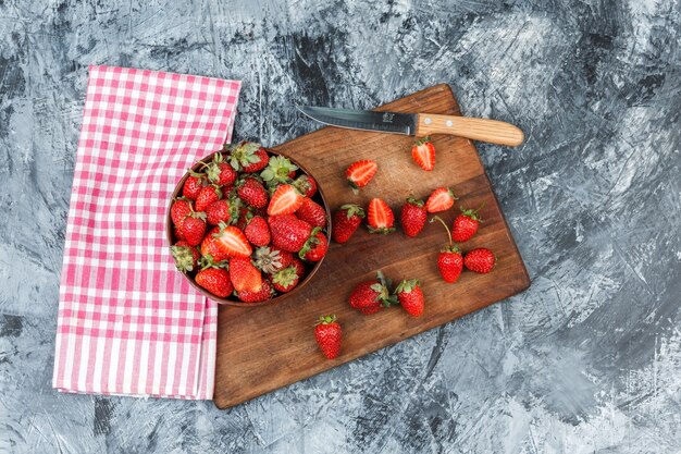 Legen Sie flach eine Schüssel Erdbeeren und ein Messer auf ein Holzschneidebrett mit roter Gingham-Tischdecke auf dunkelblauer Marmoroberfläche. horizontal