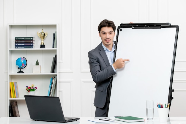 Lehrer intelligenter Ausbilder im grauen Anzug im Klassenzimmer mit Computer und Whiteboard, die auf die Tafel zeigen