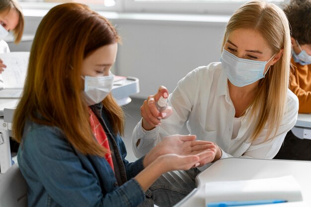 Lehrer mit medizinischer Maske, die die Hände des Schülers im Unterricht desinfiziert