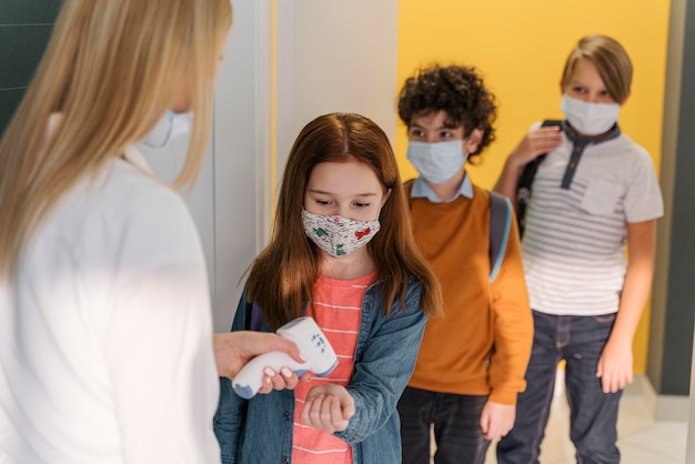 Lehrerin mit medizinischer Maske, die die Temperatur des Schülers in der Schule prüft