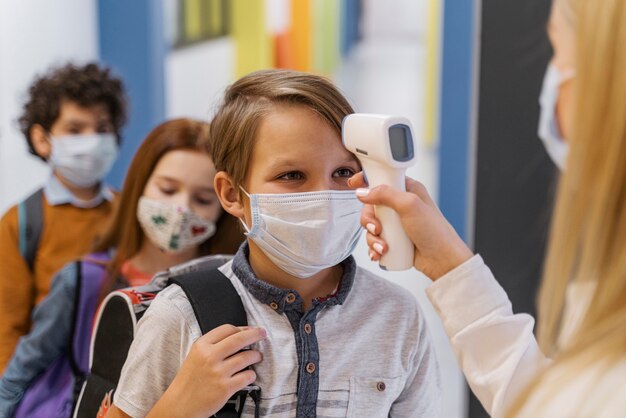 Lehrerin mit medizinischer Maske, die die Temperatur des Schülers in der Schule prüft