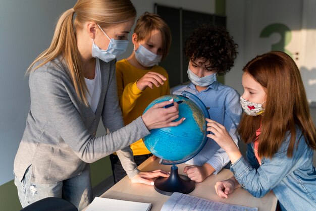 Lehrerin mit medizinischer Maske, die Geographie mit Erdkugel in der Klasse unterrichtet