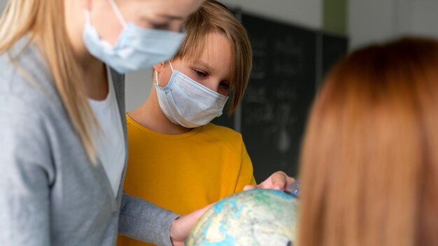 Lehrerin mit medizinischer Maske, die Geographie mit Globus im Klassenzimmer unterrichtet