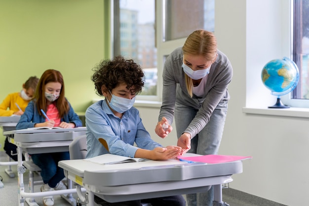 Kostenloses Foto lehrerin mit medizinischer maske, die kindern im klassenzimmer händedesinfektionsmittel gibt