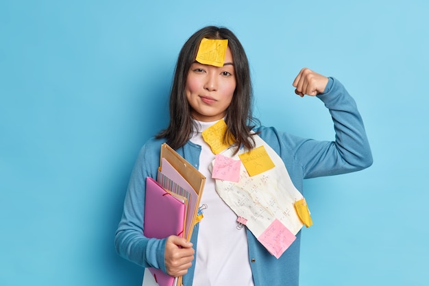 Kostenloses Foto leistungsstarke studentin hebt den arm und zeigt, dass sich die muskeln nach der arbeit an diplompapier sicher fühlen. sie trägt aufkleber auf der stirn und hält ordner.